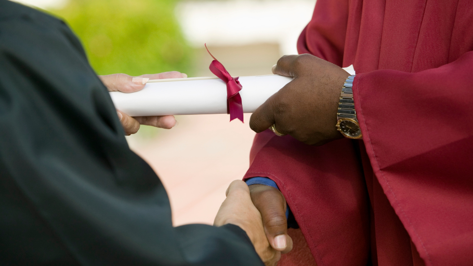 In this image, two individuals are shaking hands, and one individual is handing over a diploma to the other individual who is dressed in a graduation gown